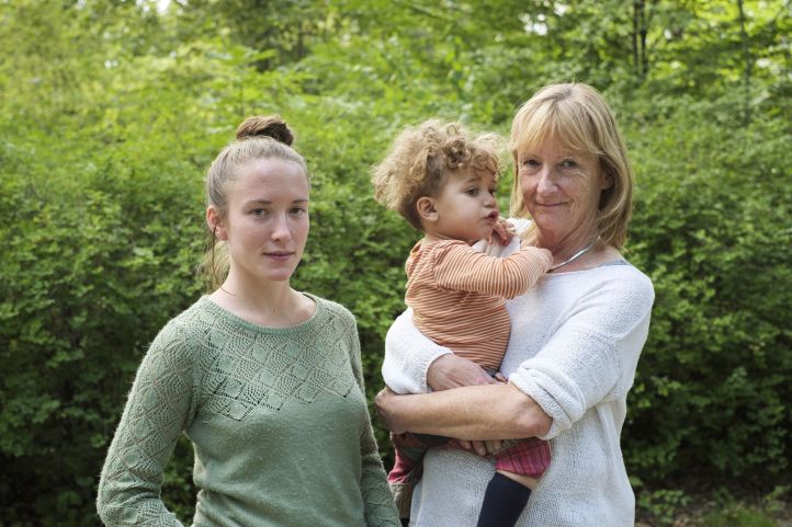 Eine junge Frau steht lächelnd neben einer etwas älteren Frau, die freundlich einen kleinen Jungen auf dem Arm hält