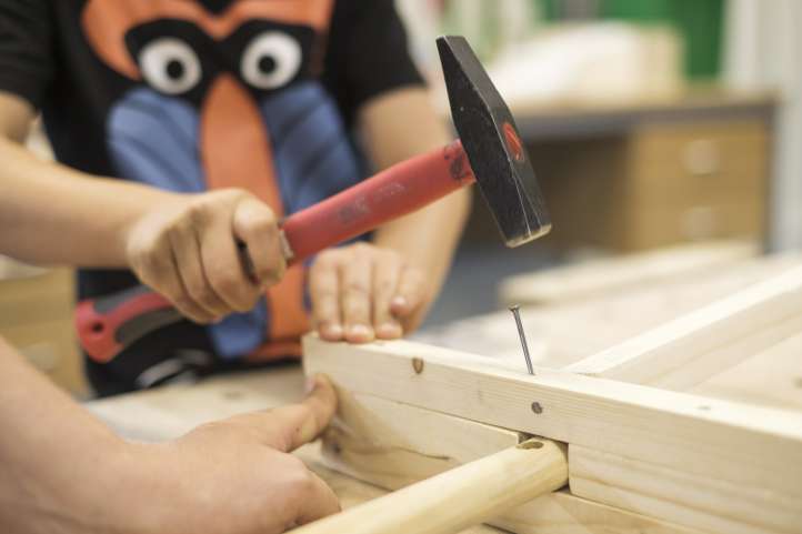 Die Hand eines Erwachsenen hält ein Holzstück, in das die Hand eines Kindes mit einem Hammer einen Nagel schlägt