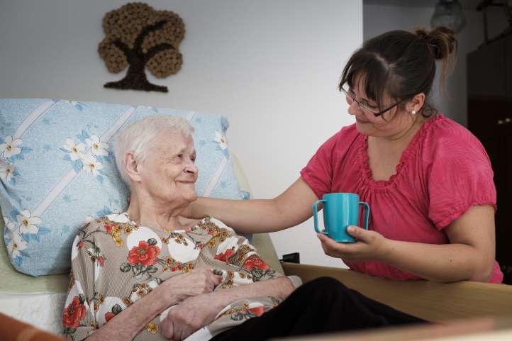 Alte Dame im Bett trinkt Kaffee. Ein Altenpflegerin reicht an.