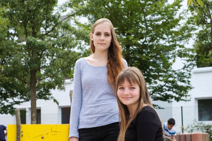 zwei junge Frauen Mädchen sitzen in einem Sandkasten umgeben von Kindern und lächeln in die Kamera