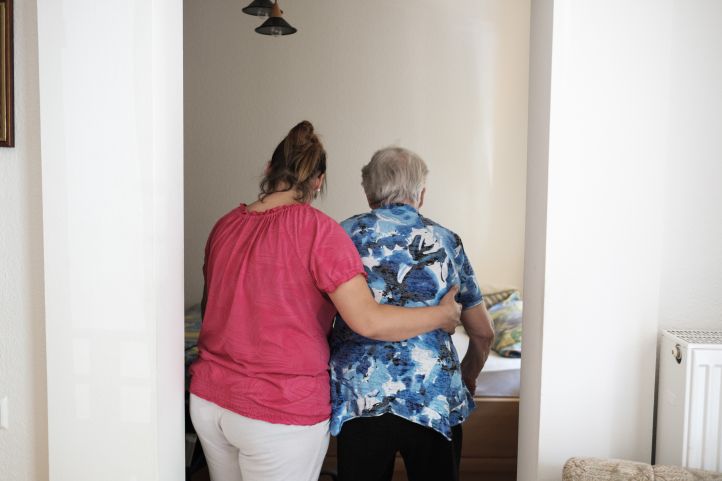 Eine Frau legt den Arm um eine Seniorin, altere Frau, und führt sie zum Bett