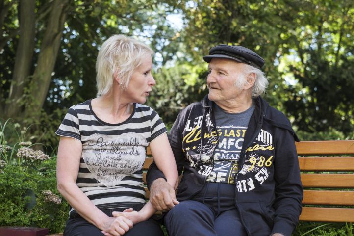 Eine Frau, wahrscheinlich eine Pflegerin, und ein älterer Mann, ein Senior mit Schiebermütze, sitzen Arm in Arm auf einer Bank im Park und schauen sich an